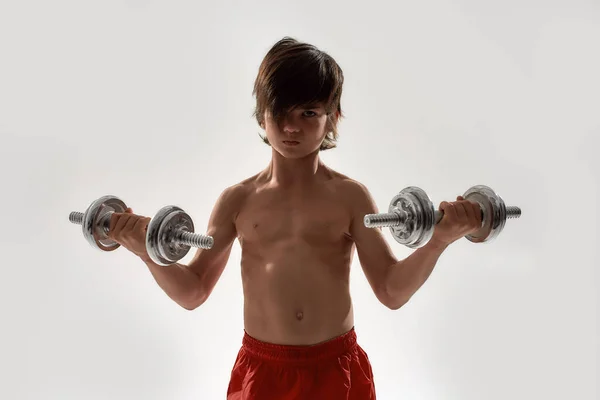 Piccolo bambino ragazzo sportivo con il corpo muscolare guardando focalizzata sulla fotocamera, sollevamento pesi mentre in piedi isolato su sfondo bianco — Foto Stock