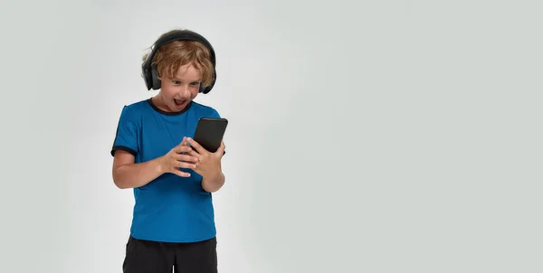 Retrato de niño pequeño deportivo en auriculares mirando emocionado en el teléfono inteligente en sus manos mientras posa, de pie aislado sobre fondo blanco — Foto de Stock