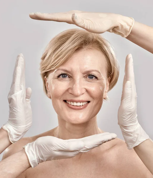 Gros plan portrait d'une jolie femme d'âge moyen souriant à la caméra. Esthéticienne créant un cadre autour du visage féminin avec les mains dans des gants isolés sur fond gris — Photo