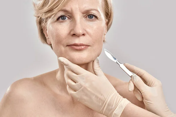 Close-up portret van aantrekkelijke vrouw van middelbare leeftijd op zoek naar camera en de medische scalpel in artsen handen geïsoleerd over grijze achtergrond — Stockfoto