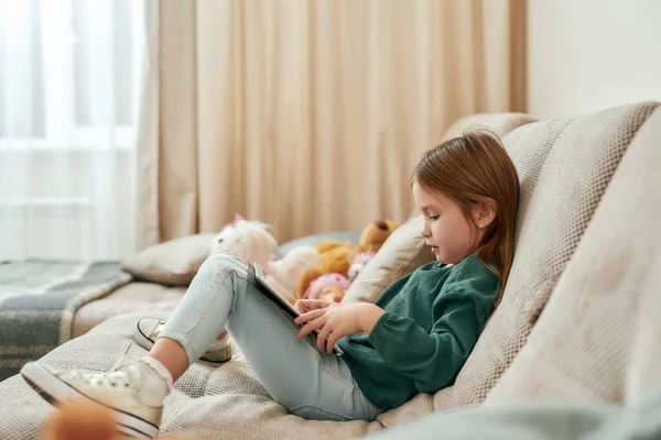 Uma menina bonita pequena está em um processo de jogar jogos em um tablet enquanto sentado em um sofá — Fotografia de Stock