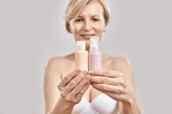 Close up of hands of middle aged woman holding two bottles of different cosmetic skincare products and comparing, choosing what to apply isolated over grey background — Stock Photo, Image