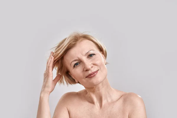 Portret van een mooie vrouw van middelbare leeftijd die naar de camera kijkt, haar haar aanpast, geïsoleerd poseert tegen een grijze achtergrond. Schoonheid, huidverzorging concept — Stockfoto