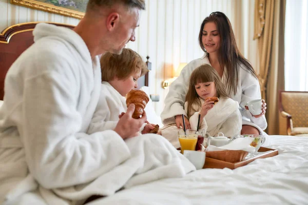 Relájate y siente paz. Primer plano de los padres y dos niños en batas blancas desayunando en la cama, comiendo y bebiendo en la lujosa habitación del hotel. Familia, resort, concepto de servicio de habitaciones — Foto de Stock