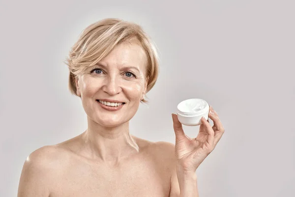 Portrait of beautiful middle aged woman smiling at camera, holding moisturizing facial cream while posing isolated over grey background — Stock Photo, Image