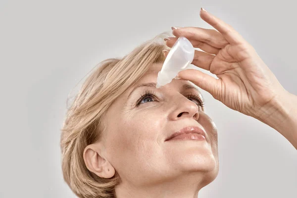 Primer plano retrato de mujer atractiva de mediana edad aplicando gotas para los ojos, de pie aislado sobre fondo gris —  Fotos de Stock