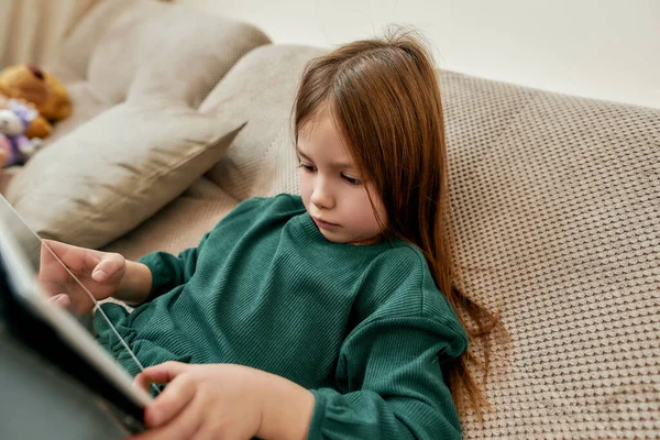 Uma menina jogando videogames em seu tablet enquanto está sentada descansando em um sofá — Fotografia de Stock