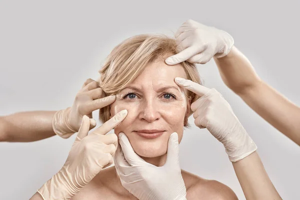 Retrato de beleza de mulher de meia-idade sorrindo para a câmera. Mãos de esteticista em luvas verificando a pele feminina do rosto isolado contra fundo cinza — Fotografia de Stock