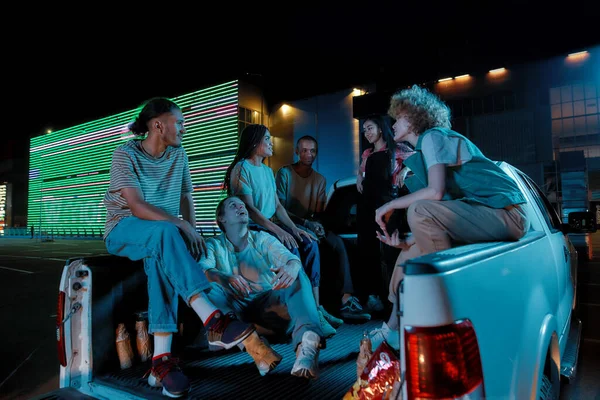 Skupina mladých dobře oblečených přátel různých národností sedí v pick-up kufru před sebou venku na parkovišti mluví s velkou LED obrazovkou za — Stock fotografie