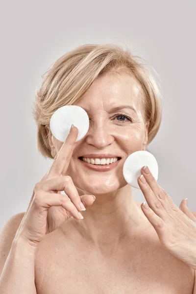 Beautiful middle aged woman with flawless skin smiling at camera while holding cotton pads near her face, posing isolated over grey background — Stock Photo, Image