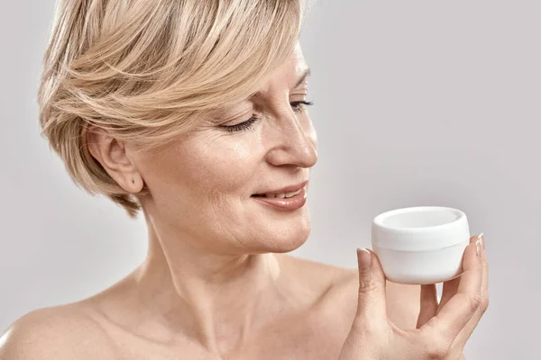 Close up portrait of beautiful middle aged woman holding, looking at moisturizing cream jar while posing isolated over grey background — Stock Photo, Image