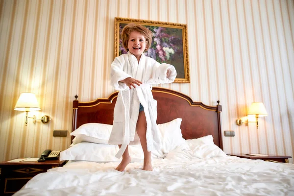 Estou a manter-te em movimento. Menino alegre em roupão branco está se divertindo, pulando na cama branca em um quarto de hotel. O garoto ri e salta — Fotografia de Stock