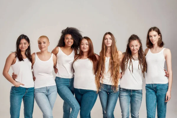 Group of beautiful diverse young women wearing white shirt and denim jeans looking at camera while posing together isolated over grey background — Stock Photo, Image