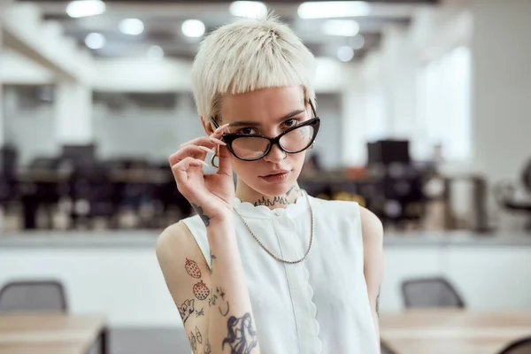 Genç ve başarılı. Kendine güvenen sarışın dövmeli bir iş kadınının portresi. Gözlüğünü ayarlıyor ve modern çalışma alanında kameraya bakıyor. — Stok fotoğraf