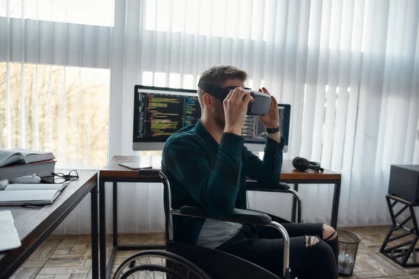 Jonge mannelijke webontwikkelaar in een rolstoel met virtual reality bril tijdens het werken op zijn werkplek in modern kantoor. Meerdere computerschermen met programmacode — Stockfoto