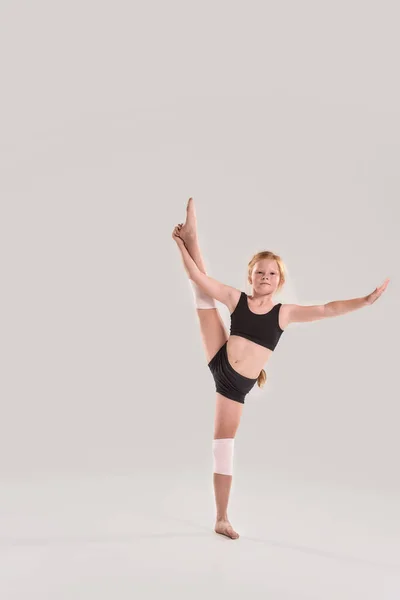 Full length shot of cute little redhead girl gymnast looking at camera, raising one leg with her hand, showing flexibility isolated over grey background — Stock Photo, Image