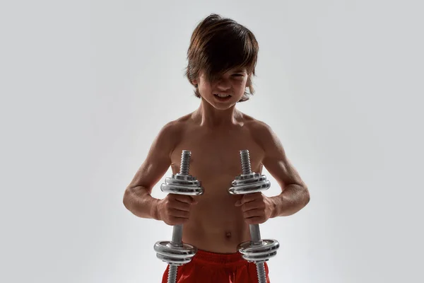 Pequena criança menino esportivo com corpo muscular olhando emocional durante o exercício, levantando pesos, de pé isolado sobre fundo cinza — Fotografia de Stock