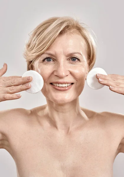 Beautiful middle aged woman with flawless skin smiling at camera while holding cotton pads near her face, posing isolated over grey background — Stock Photo, Image