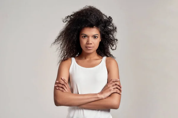 Ritratto di giovane donna afroamericana con i capelli ricci che indossa camicia bianca guardando la macchina fotografica mentre in piedi con le braccia incrociate isolato su sfondo grigio — Foto Stock