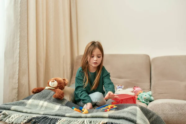 Roztomilá holčička vybírá hračku stetoskop z kostkované sedí na pohovce v blízkosti plyšového medvídka v světlé místnosti — Stock fotografie