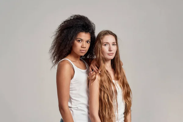 Ritratto di due giovani donne diverse che indossano camicie bianche guardando la macchina fotografica mentre posano insieme isolate su sfondo grigio — Foto Stock
