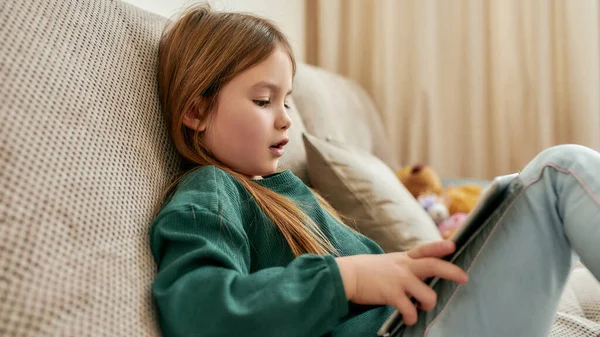 Uma menina pequena jogando jogos em seu tablet enquanto sentada em um sofá — Fotografia de Stock