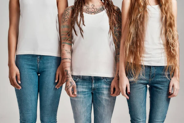 Colpo ritagliato di tre giovani donne diverse che indossano camicie bianche e jeans di jeans in piedi insieme isolati su sfondo grigio — Foto Stock