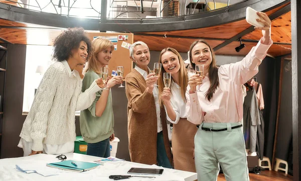 Cinco mujeres bien vestidas de diferentes edades y razas con copas de champán de pie juntas sonriendo y esperando una selfie — Foto de Stock