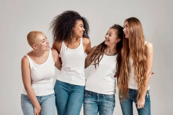 Ritratto di quattro giovani donne diverse che indossano camicie bianche e jeans di jeans che si sorridono mentre posano, in piedi insieme isolate su sfondo grigio — Foto Stock