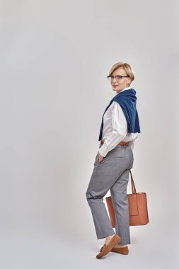 İş elbisesi ve gözlük takmış zarif orta yaşlı bir kadının el çantasıyla poz verirken gri arka planda tek başına dikilip kameraya baktığı uzun bir kare.