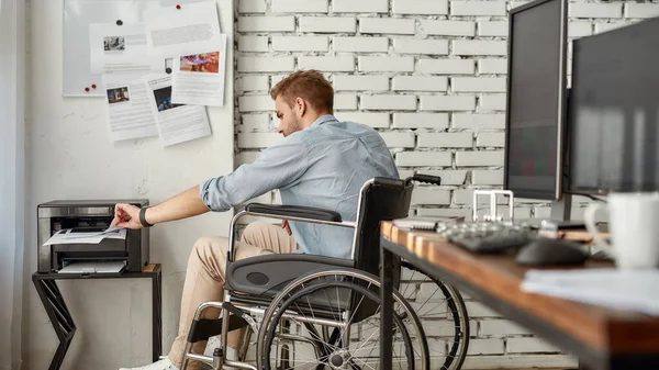 Kantoorroutine. Zijaanzicht van jonge mannelijke kantoormedewerker in een rolstoel die een dossier drukt op zijn werkplek in het heldere moderne kantoor. Gehandicapten en handicaps — Stockfoto