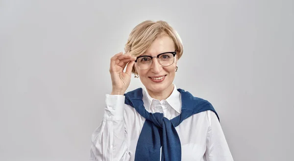 Retrato de una elegante mujer caucásica de mediana edad con atuendo de negocios ajustando sus gafas, sonriendo a la cámara mientras está de pie aislada sobre un fondo gris —  Fotos de Stock