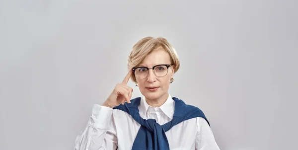 Retrato de una elegante mujer caucásica de mediana edad que usa ropa de negocios tocando sus gafas con un dedo, mirando a la cámara mientras está de pie aislada sobre un fondo gris —  Fotos de Stock