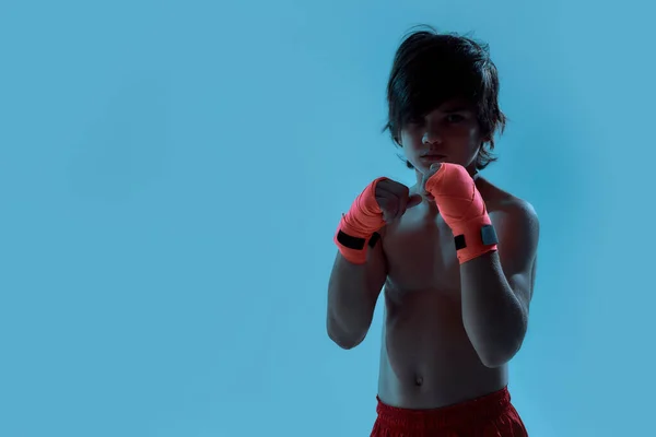 Silhouetted shot of little boy boxer in shorts with boxing bandage on hands, looking at camera ready to fight while posing, standing isolated over blue background — Stock Photo, Image