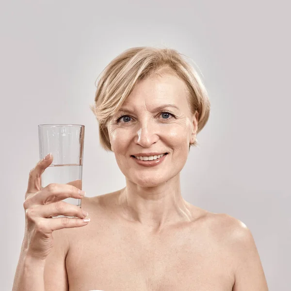 Retrato de mulher de meia-idade atraente sorrindo para a câmera enquanto segurava vidro de água na mão, posando isolado sobre fundo cinza — Fotografia de Stock