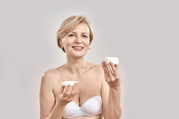 Portrait of beautiful middle aged woman smiling at camera, holding moisturizing facial cream while posing isolated over grey background — Stock Photo, Image