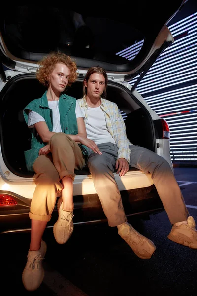 Dva mladí běžně oblečení bílí přátelé různého pohlaví hledí do kamery pózovat, zatímco sedí v otevřeném kufru auta venku na parkovišti s LED obrazovkou za — Stock fotografie