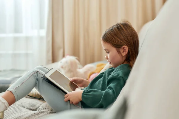 A cute small girl playing videogames on a tablet while sitting on a sofa — Stock Photo, Image