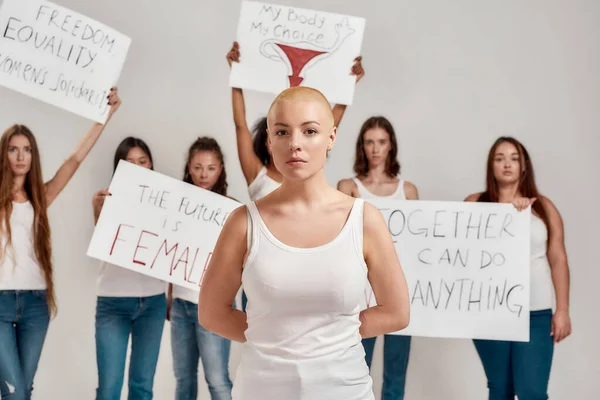 Молодая белая женщина с бритой головой в белой рубашке, смотрящая в камеру. Группа различных женщин держит плакаты протеста за женскую власть и права на сером фоне — стоковое фото