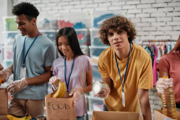 Young male volunteer looking at camera while packing food and drinks donation into paper bags and box, Small group of people working in charitable foundation