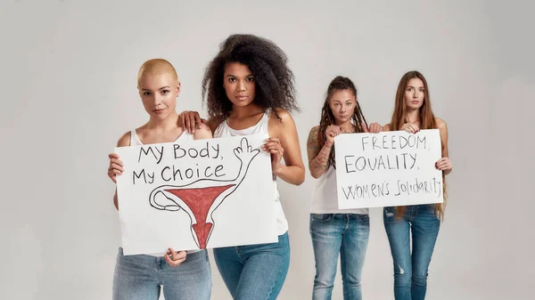4人の多様な女性がカメラを見ながら、灰色の背景にバナーを手に立っている。フェミニズムと平等の概念 — ストック写真