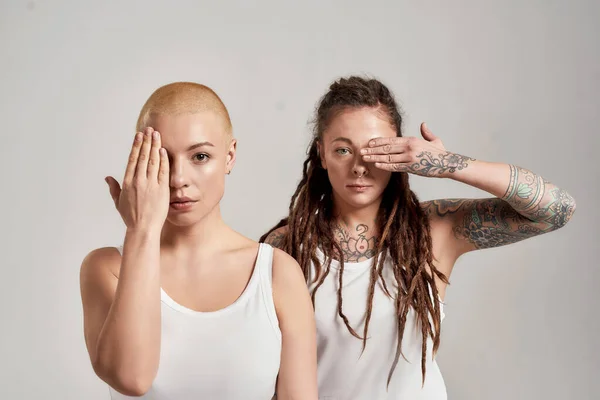 Ritratto di due diverse giovani donne caucasiche che indossano camicia bianca, guardando la macchina fotografica, coprendo metà del viso con la mano mentre posano insieme isolate su sfondo grigio — Foto Stock