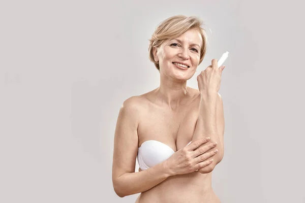 Attractive middle aged woman smiling at camera, applying exfoliating moisturizing cream on dry elbow skin while posing isolated over grey background — Stock Photo, Image