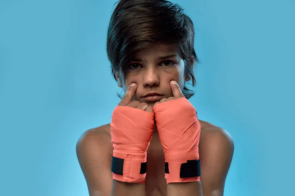 Retrato de cerca del pequeño boxeador con vendaje de boxeo en las manos, mirando a la cámara, tomándose de las manos cerca de su cara mientras posa aislado sobre fondo azul — Foto de Stock