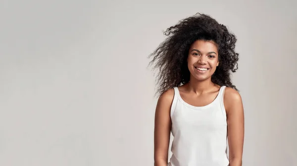 Ritratto di giovane donna afroamericana con i capelli ricci che indossa camicia bianca sorridente alla macchina fotografica mentre in piedi isolato su sfondo grigio — Foto Stock