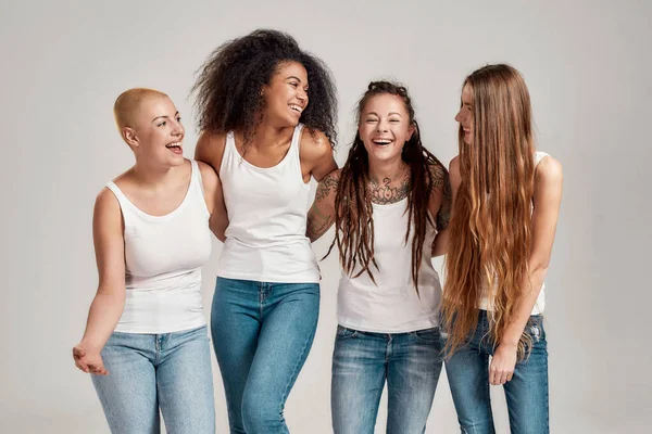 Portrait of four young diverse women wearing white shirts and denim jeans laughing together while posing, standing isolated over grey background