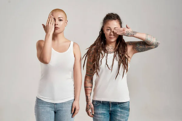 Due diverse giovani donne caucasiche che indossano camicia bianca, guardando la fotocamera, coprendo metà del viso con la mano mentre posano insieme isolati su sfondo grigio — Foto Stock