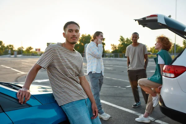 An ordinary dressed dark-skinned latina man with a piercing looking into a camera leaning against a car standing outside on a parking site with his friends on a background