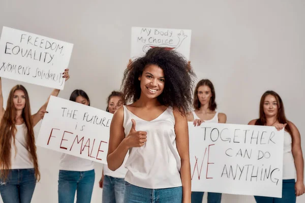 白いシャツに身を包んだ若いアフリカ系アメリカ人女性がカメラを見て微笑んだ。背景に女性の力と権利のための抗議のバナーを保持する多様な女性のグループ — ストック写真