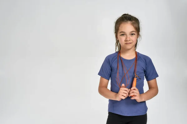 Маленькая спортивная девочка в спортивной одежде смотрит в камеру, стоит с скакалкой, позируя изолированно на белом фоне — стоковое фото